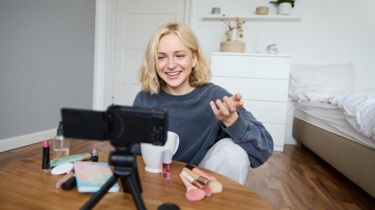 Junge Creatorin filmt Vlog mit Make-up-Produkten in gemütlichem Schlafzimmer.